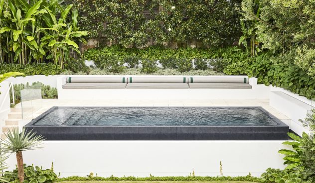 A large sparkling pool with built in day bed sits elevated at the rear of the property surrounded by large trees and plants including magnolia, heliconia and raphiolepsis. The pool floats above a large green level lawn.